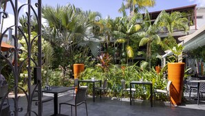 Un patio al aire libre con mesas y sillas, rodeado de exuberante follaje tropical y una cerca de metal.