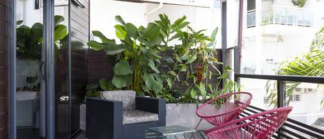 Un balcón con una mesa de vidrio, dos sillas rojas y una jardinera con plantas verdes grandes.