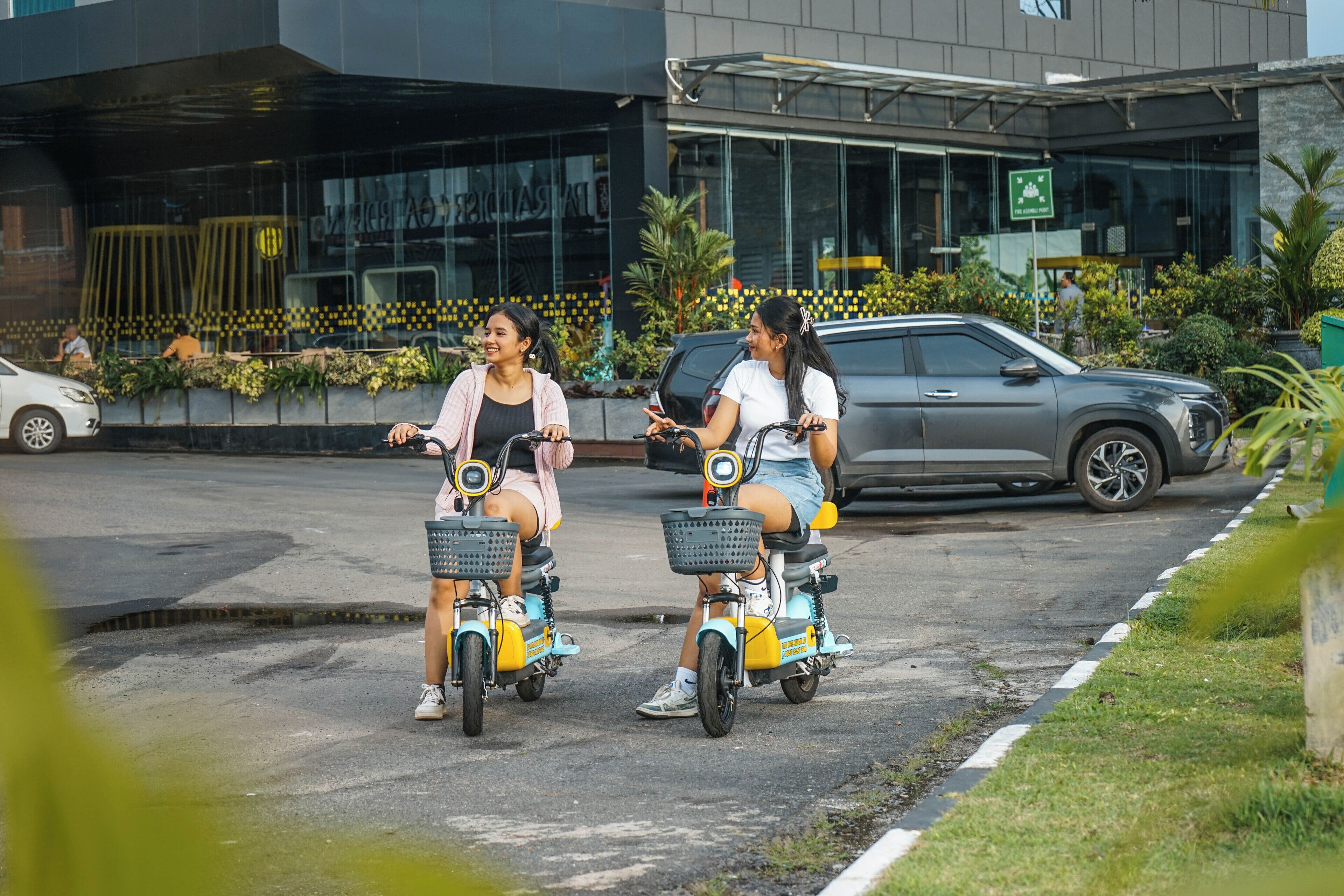 Two people riding electric scooters on a street with a modern building in the background.