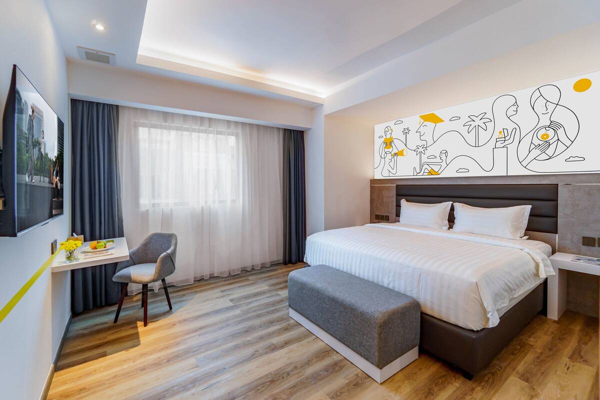 A modern hotel room with a large bed, a desk with a chair, and a large window with curtains.
