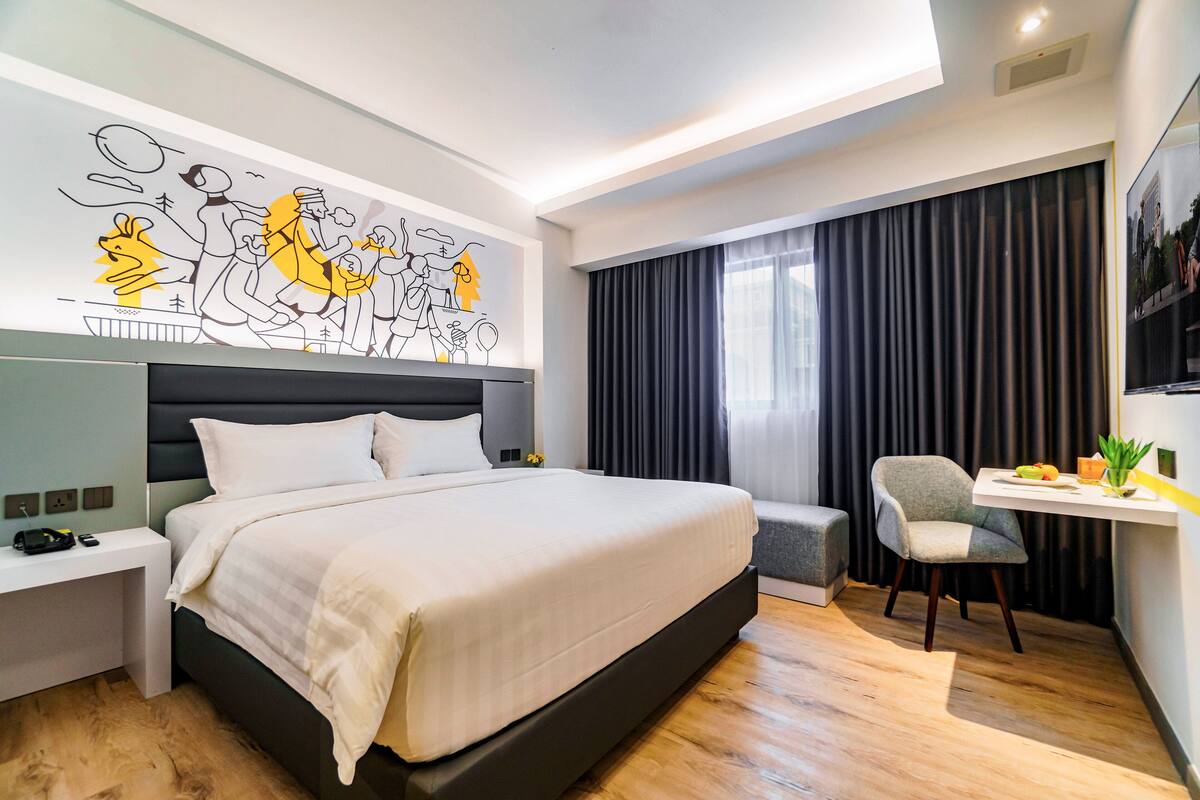 A modern hotel room with a large bed, a desk with a chair, a wall mural, and a large window with curtains.