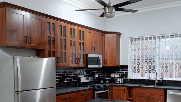 A kitchen with wooden cabinets, a stainless steel refrigerator, a microwave, a stove, and a window with a lattice design. - 4 Bedrooms Family House Sakumono (Sakumono)