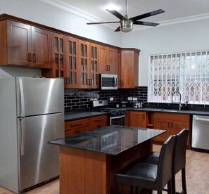 A kitchen with wooden cabinets, a stainless steel refrigerator, a microwave, a stove, and a window with a lattice design. - 4 Bedrooms Family House Sakumono (Sakumono)