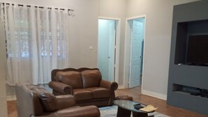 A living room with a brown leather sofa set, a glass coffee table, and a patterned rug. There is a flat-screen TV mounted on the wall. - 4 Bedrooms Family House Sakumono (Sakumono)