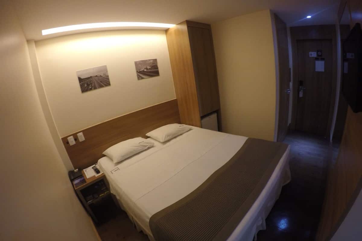 A hotel room with a bed, bedside tables, a nightstand, a wooden headboard, and a wall-mounted light fixture.