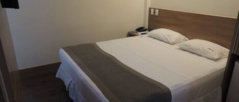 Una cama bien hecha con edredón blanco y dos almohadas, cabecera de madera, mesita de noche con teléfono y un interruptor de luz en la pared.