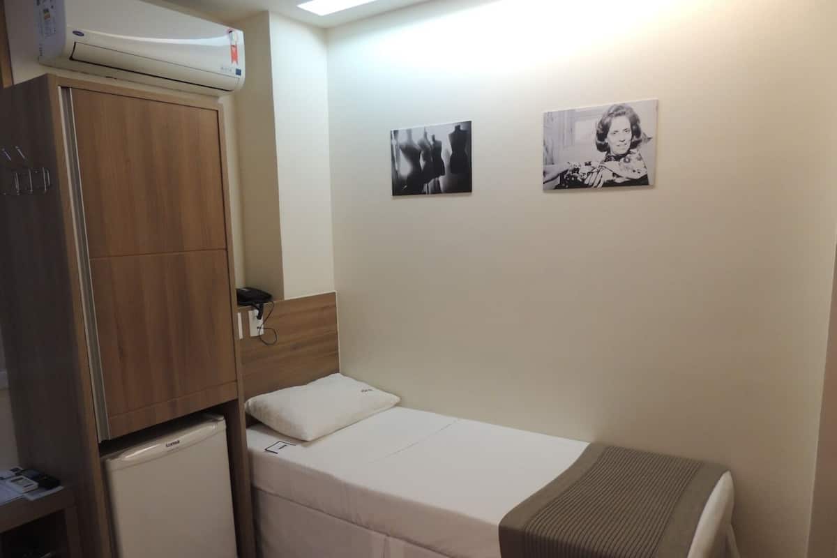 A small, compact room with a single bed, a bedside table, a wardrobe, and a small refrigerator. There are two wall-mounted pictures.