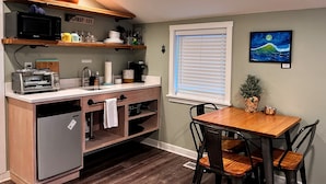 A compact kitchen with a built-in dishwasher, a microwave, and a toaster. There's a window with blinds, a small dining table with chairs, and a potted plant on the table. - Sea Glass Inn (Seaside)