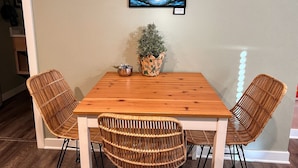A dining area with a wooden table and four wicker chairs. A framed painting of a snowy landscape hangs above the table. - Sea Glass Inn (Seaside)