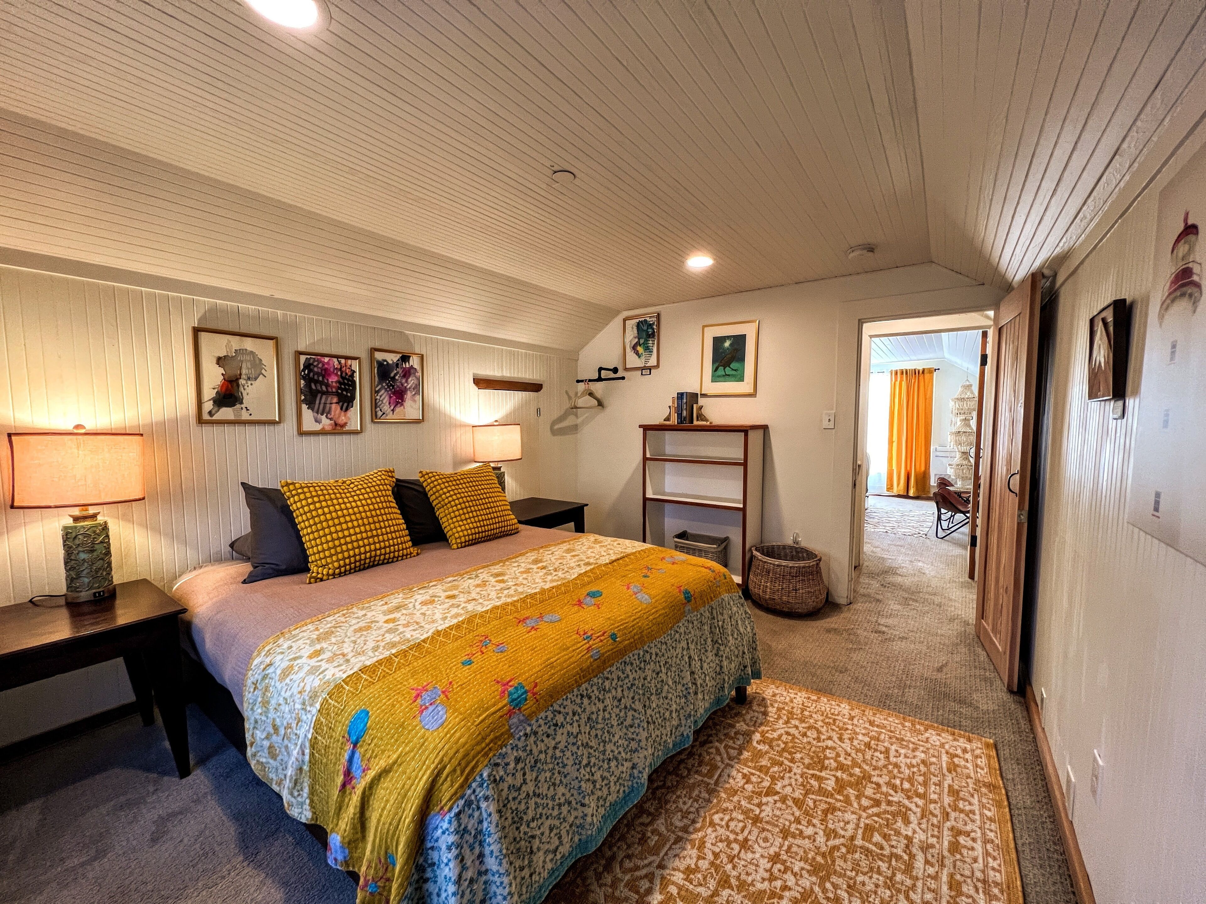Un dormitorio con cama, mesitas de noche, lámpara, estante con libros y cuadros en la pared.