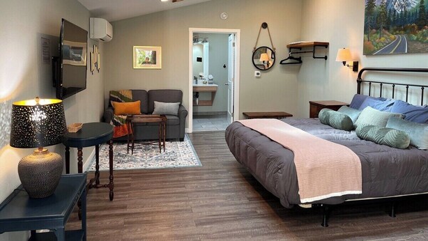 A modern bedroom with a large bed, a desk, and a sofa. There is a painting on the wall and a door leading to another room. - Sea Glass Inn (Seaside)