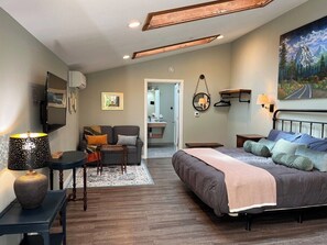 A modern bedroom with a large bed, a desk, and a sofa. There is a painting on the wall and a door leading to another room. - Sea Glass Inn (Seaside)