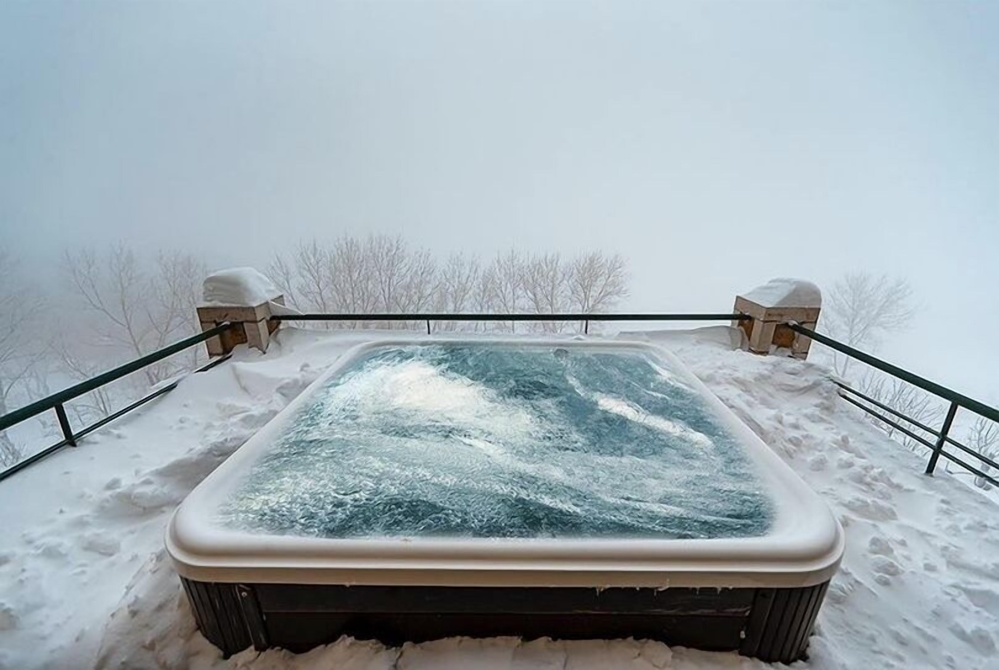 Un bain à remous enneigé sur un balcon doté d’une balustrade, entouré de neige et d’arbres sans feuilles.