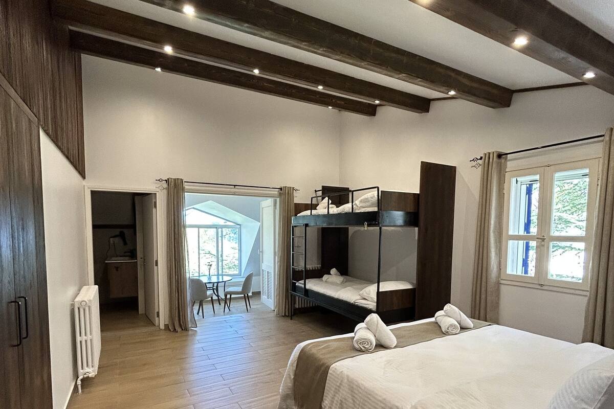 A hotel room with a large bed, a smaller bunk bed, wooden flooring, and a view of the outdoors through a window.