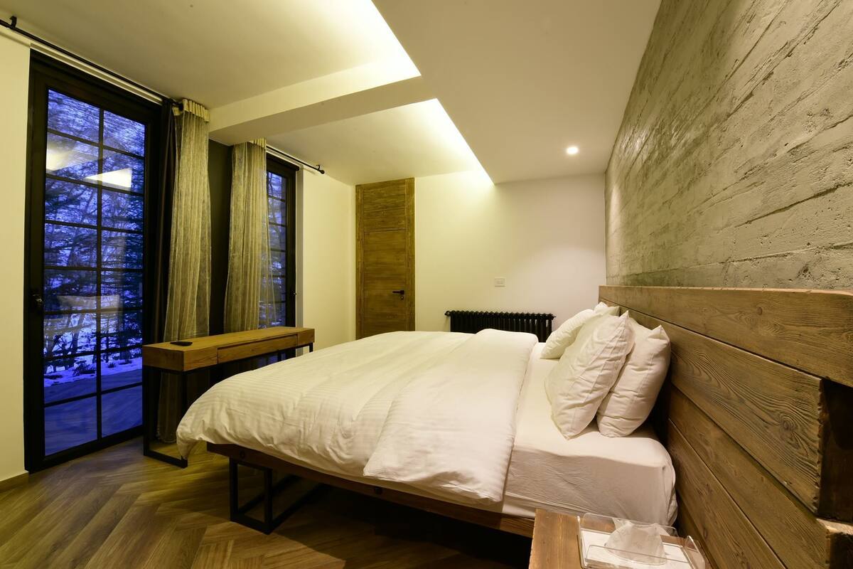 A bedroom with a wooden headboard, a large bed, a wooden bench, and a view of a snowy landscape through a window.