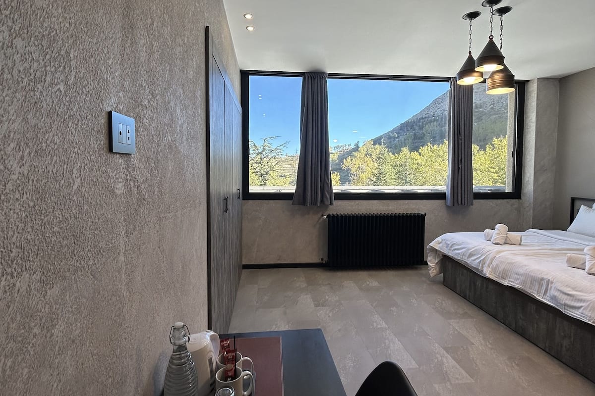 A modern hotel room with a large window, a bed, a desk with a tray of glasses and a bottle, and a radiator.