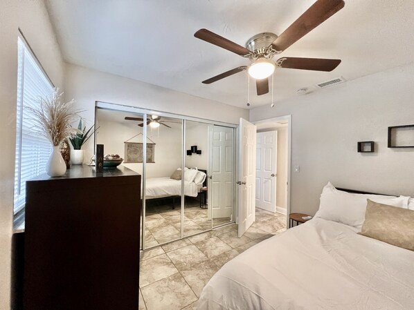 Un dormitorio con cama, ventilador de techo, cómoda, espejo y ventana con persianas.