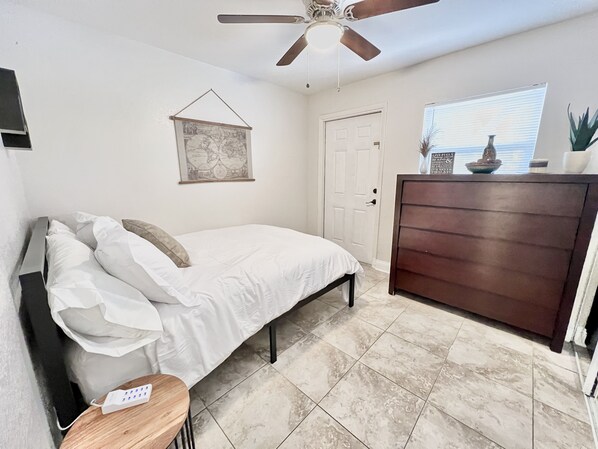 A bedroom with a bed, a wooden nightstand, a dresser, a ceiling fan, a door, a window with blinds, and a wall-mounted map.