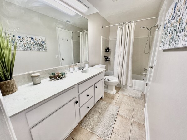 A modern bathroom with a white vanity, a large mirror, a toilet, a shower with a curtain, and a potted plant.