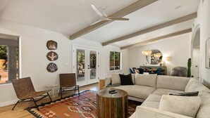 Ein geräumiges Wohnzimmer mit einer großen, beigefarbenen Eckcouch, einem Holztisch und einem farbenfrohen Teppich. Im Hintergrund sind zwei Sessel, ein Deckenventilator und ein Essbereich zu sehen.