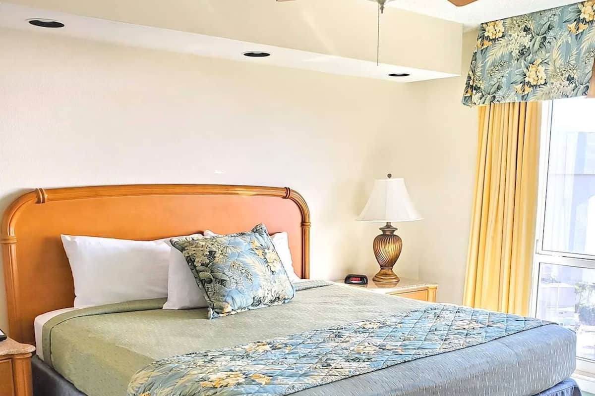 Habitación de hotel con cabecera de madera, cama con ropa de cama floral, mesita de noche con lámpara y ventana con cortinas.