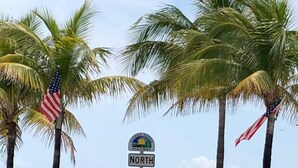 Schild am Strand mit Palmen und der US-amerikanischen Flagge, das einen Küstenort kennzeichnet.