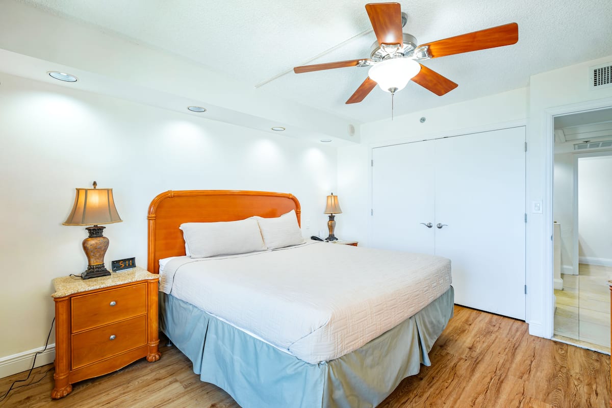 Un dormitorio con una cama de madera, dos lámparas de noche, un ventilador de techo y un armario blanco.