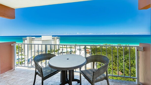 Ein Balkon mit einem runden Tisch und zwei Korbstühlen, mit Blick auf einen Strand mit klarem türkisblauem Wasser und eine entfernte Skyline.