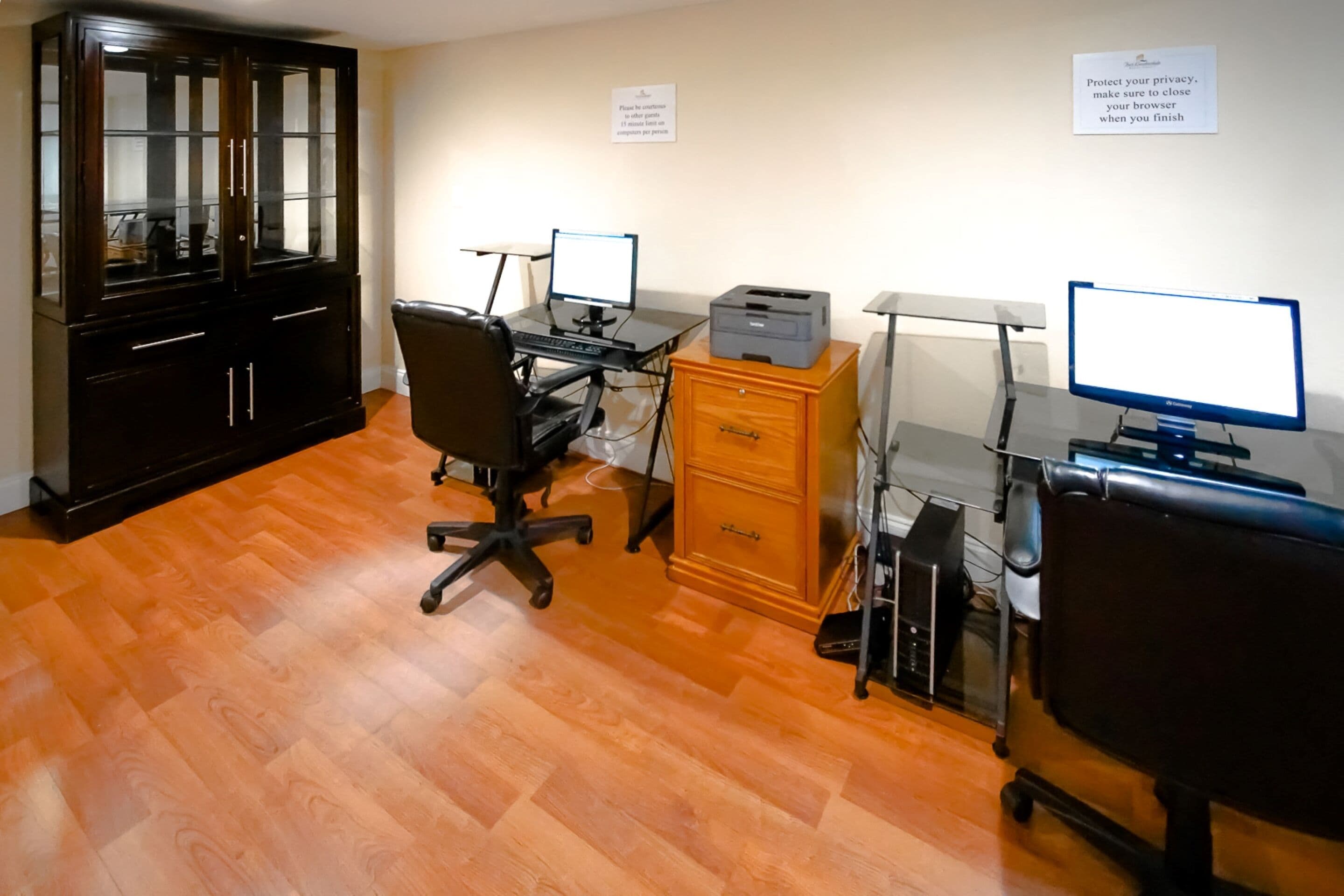 Une pièce avec un plancher en bois, une armoire noire, un bureau avec un ordinateur, une imprimante et une chaise.