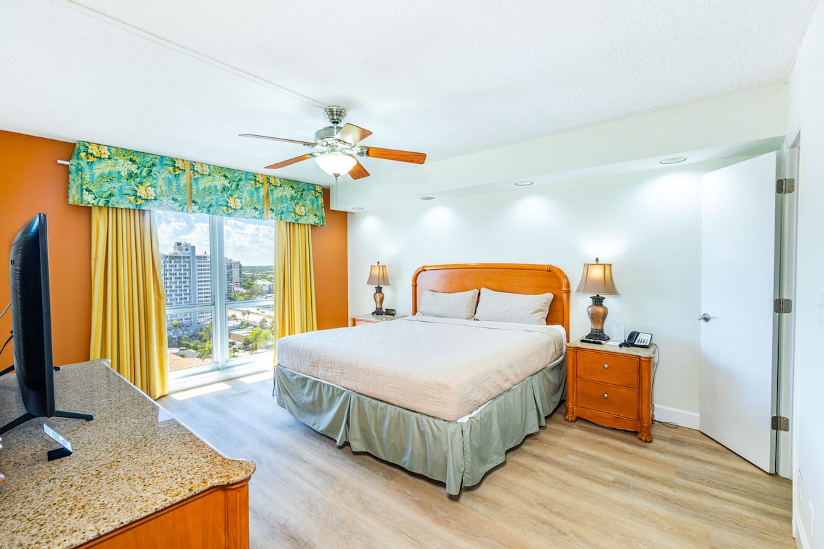 Habitación de hotel con cama, lámparas de noche, un escritorio con computadora, ventilador de techo y vista a la ciudad a través de amplias ventanas.