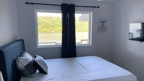 Ein Schlafzimmer mit einem Bett, einem Fenster mit Blick auf Grünflächen, einem Deckenventilator und einem an der Wand befestigten Regal.