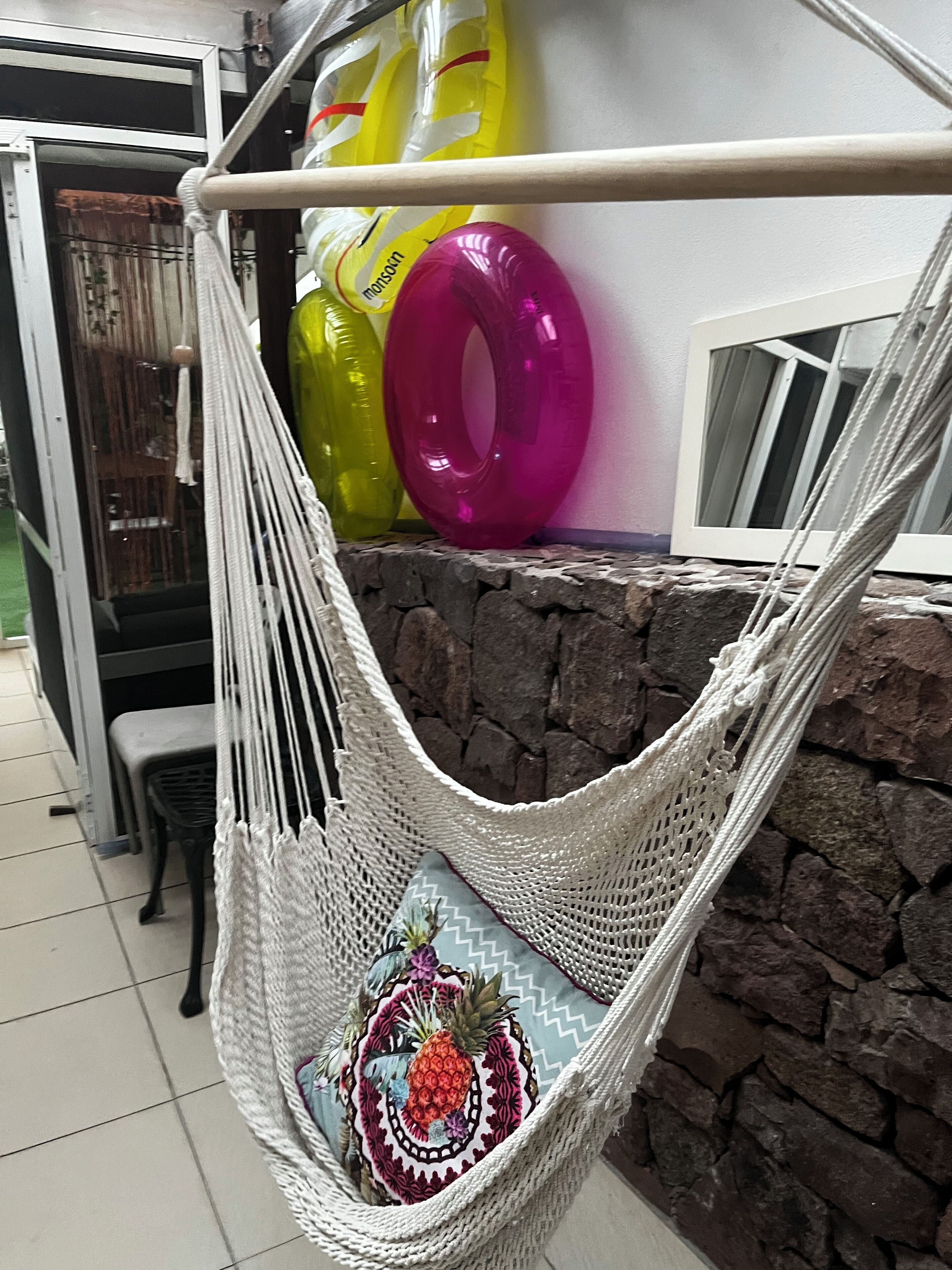Una silla hamaca con un cojín decorativo, una pared de piedra y una ventana con persianas.