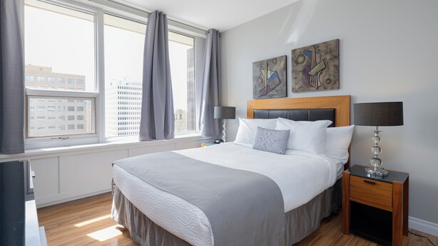 Une chambre d’hôtel moderne dotée d’un grand lit, d’une tête de lit en bois, de deux tables de chevet avec des lampes, d’une vue sur la ville grâce à de grandes fenêtres et d’œuvres d’art abstraites accrochées aux murs.
