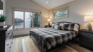 A bedroom with a large window, a bed with a checkered bedspread, a nightstand with a lamp, and a dresser with drawers. - New House on Big Sebago w/ Private beach & dock! (Casco)