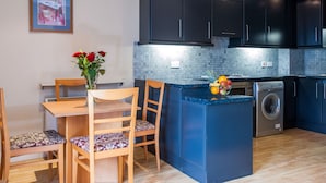 Une cuisine moderne avec des meubles de couleur sombre, un crédence en carreaux bleus, un lave-linge encastré et un coin repas avec une table et des chaises en bois.