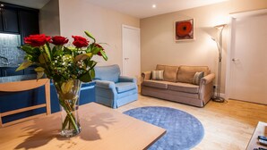 Un salon avec un fauteuil bleu, un canapé beige, une table en bois ornée d’un vase de roses rouges et un lampadaire.
