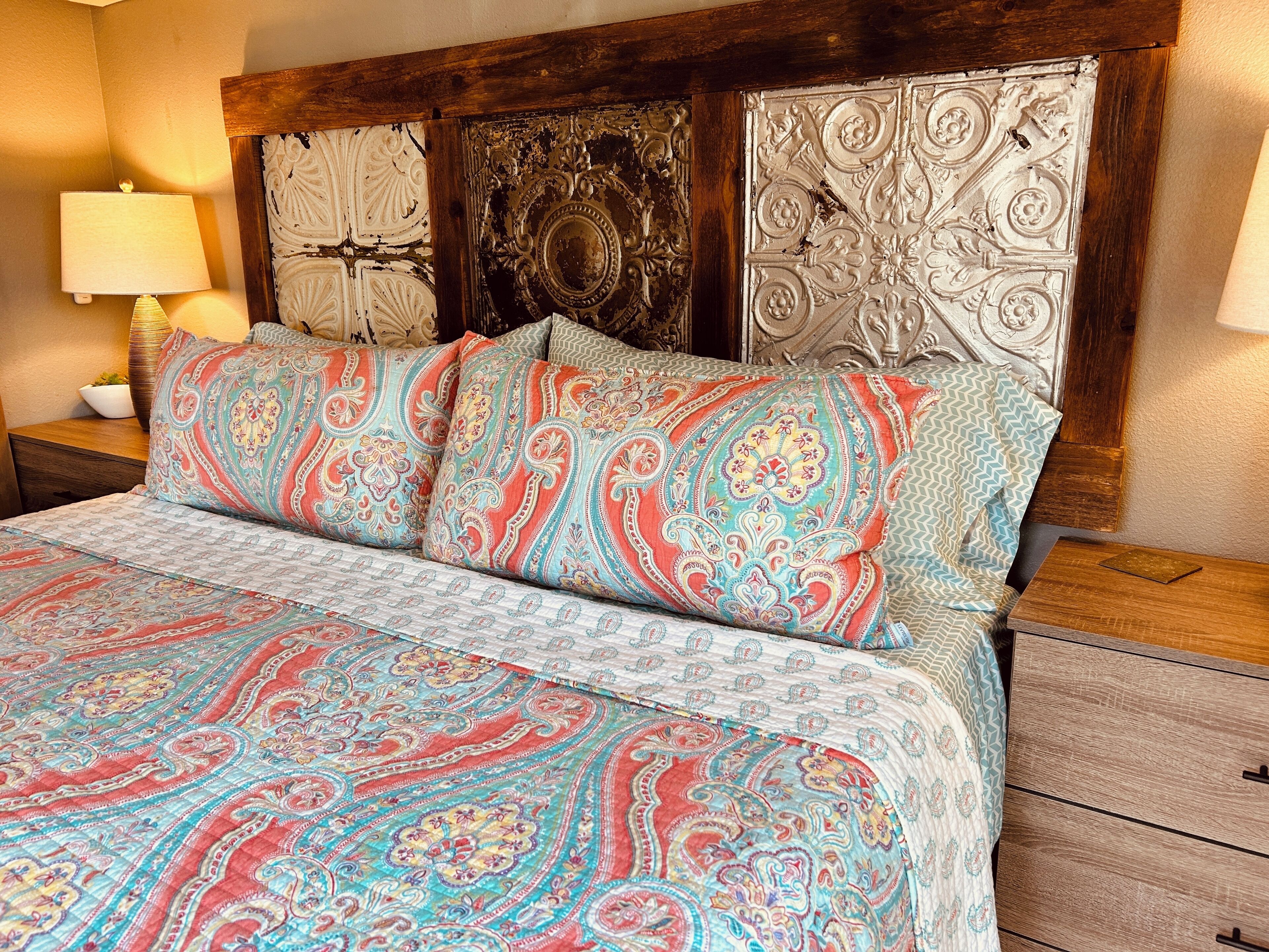 Una cama con una colcha estampada y varios almohadones, una mesita de noche de madera con una lámpara, y un cabecero decorativo con diseños intrincados.