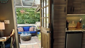 Et soveværelse med en stor seng, en trædør, et tekøkken og udsigt til en udendørs patio med sofa og planter.