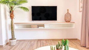 Ett vardagsrum med en tv monterad på en vägghylla, en krukväxt i form av en palm och flera olika sorters kaktusar på ett runt bord.