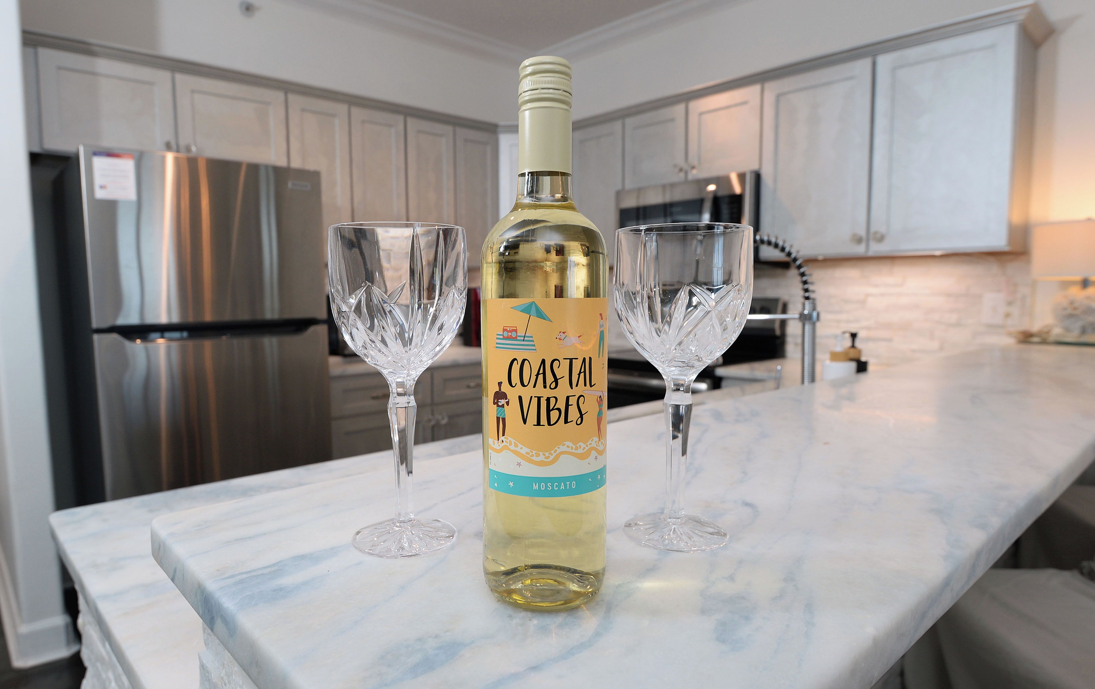 Una ampolla de vi Moscató Coastal Vibes sobre un taulell de marbre amb dues copes de vi.