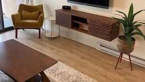 Un salon moderne avec un téléviseur à écran plat fixé au mur, une table basse en bois, un tapis à motifs, un fauteuil jaune et une plante en pot.
