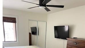 Une chambre avec deux lits, une commode en bois, un ventilateur de plafond, une télévision et une fenêtre avec des stores.