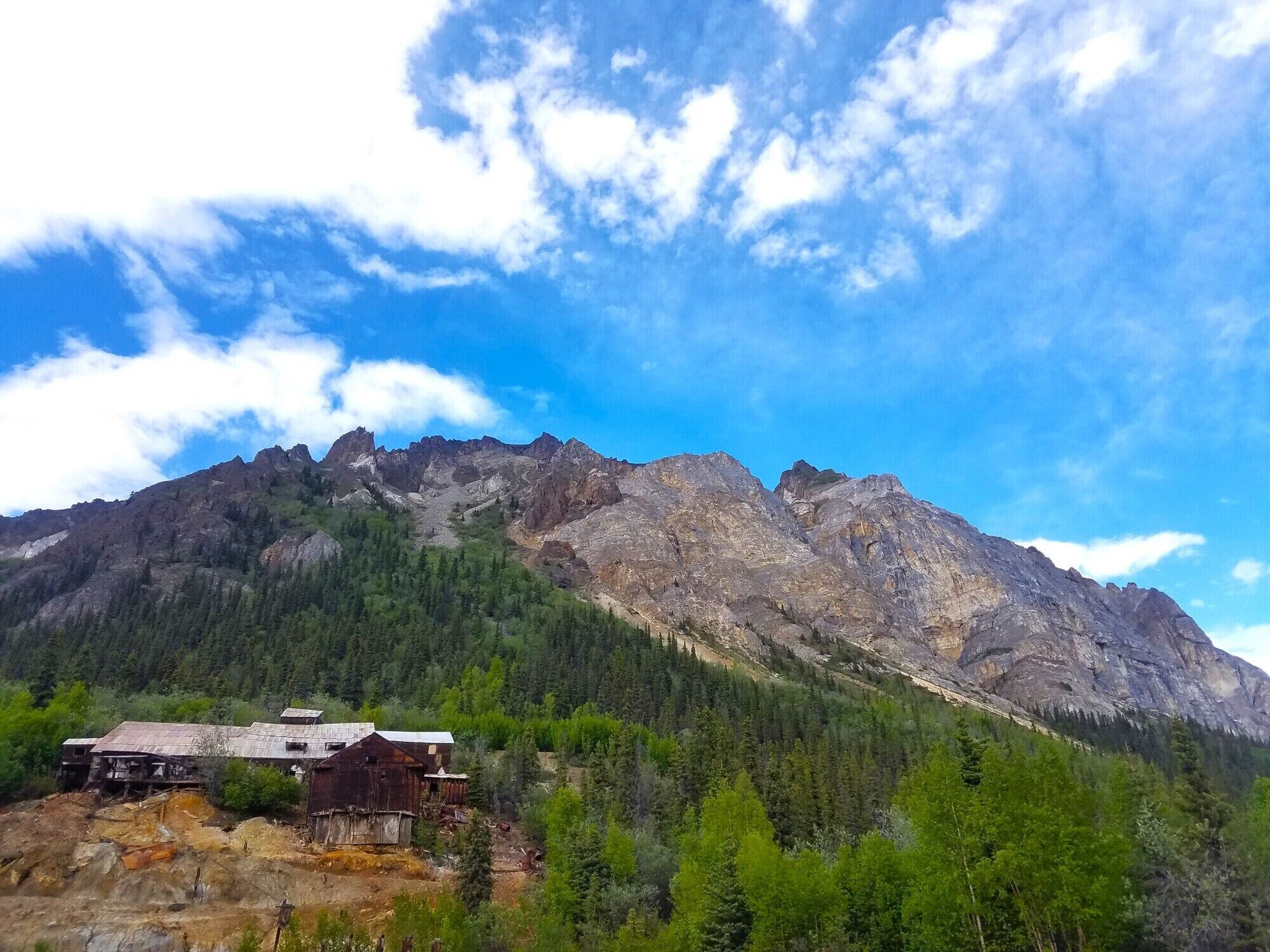 Paesaggio montano con un edificio di legno rustico, immerso in una fitta foresta e terreno roccioso.