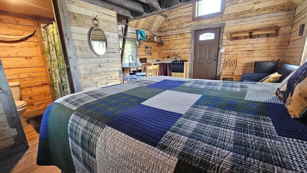 A cozy log cabin bedroom with a large bed, wooden walls, a small kitchen area, and a bathroom.