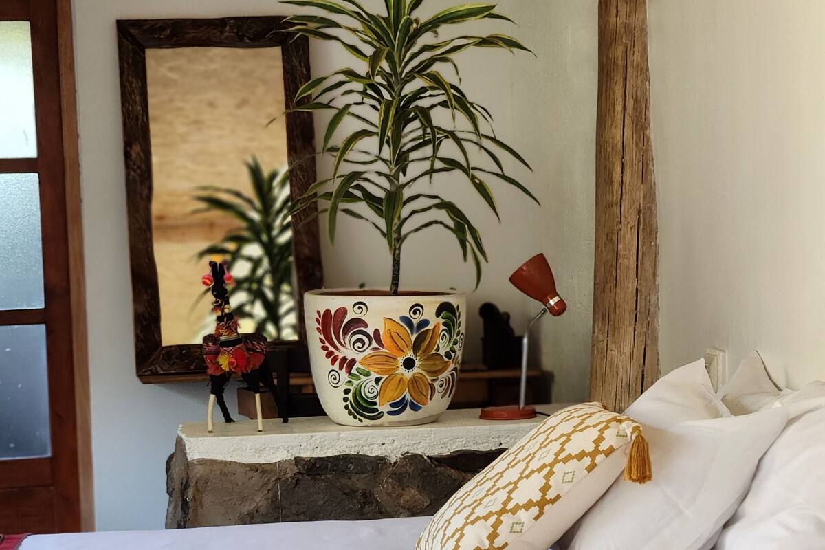 A bedroom with a bed, pillows, a decorative plant in a colorful pot, a mirror, and a wooden beam.