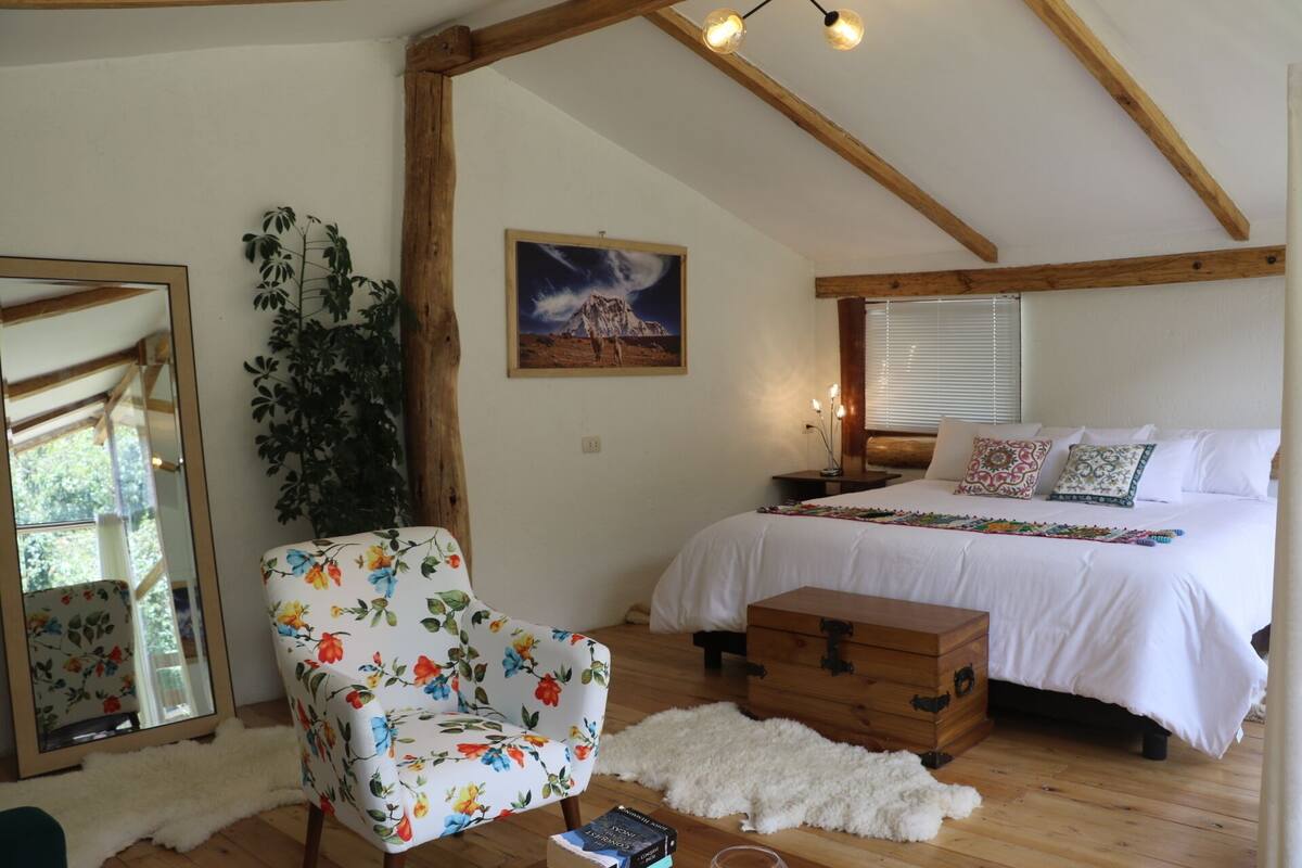 A bedroom with a wooden ceiling, a bed with white bedding, a floral armchair, a wooden chest, and a mirror.