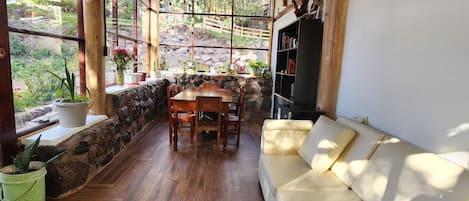 Una habitación acogedora con techo de madera, paredes de piedra, una mesa de madera con sillas, un sofá color beige y una estantería.