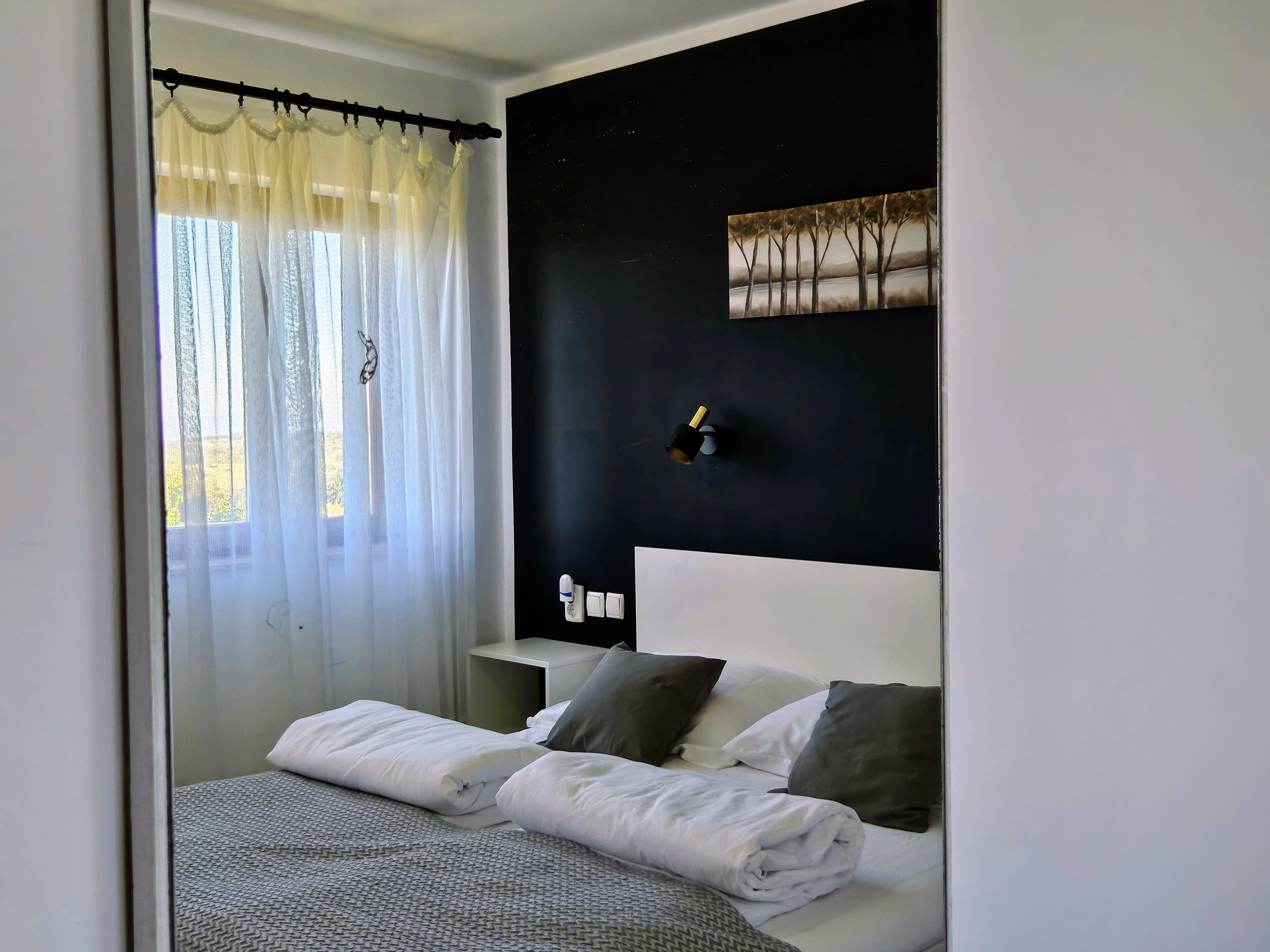 A neatly made bed with white and gray bedding, a black wall with a painting, and a window with sheer curtains.