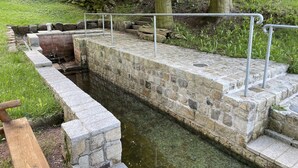 Ein steinerner Springbrunnen mit Stufen, die in einen Teich führen, umgeben von Grünpflanzen und einer Holzkonstruktion darüber.