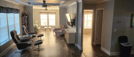A spacious living room with a large window, a ceiling fan, and a flat-screen TV mounted on the wall.